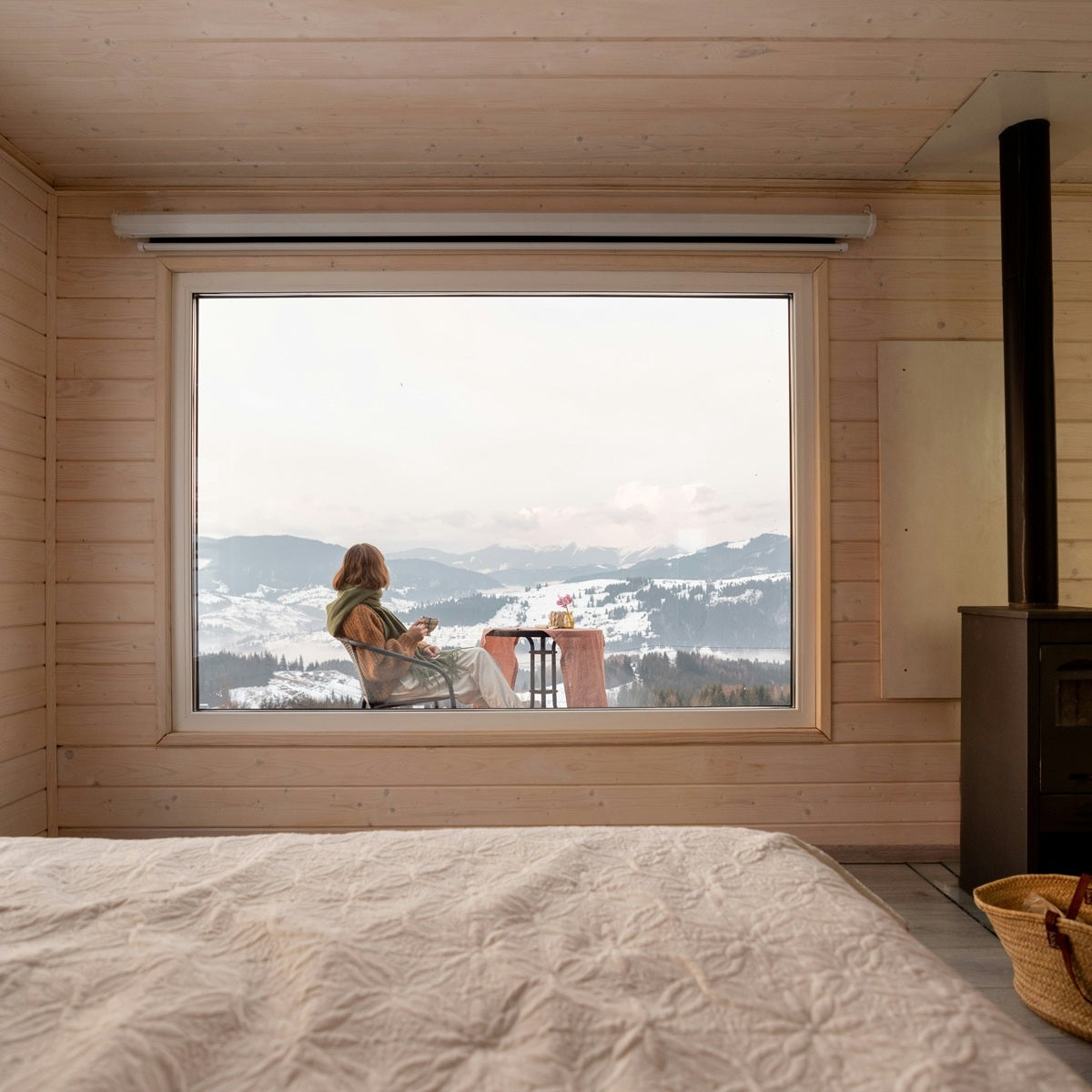 Grosses Fenster in einer Berghuette, Frau sitzt auf der Terrasse, Blick auf verschneite Berge, Holzdecke und Wand