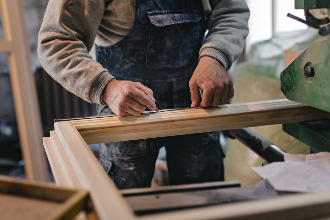 Hand eines Arbeiters misst Holzrahmen eines Fensters, Massband und Bleistift sichtbar, Werkstattumgebung