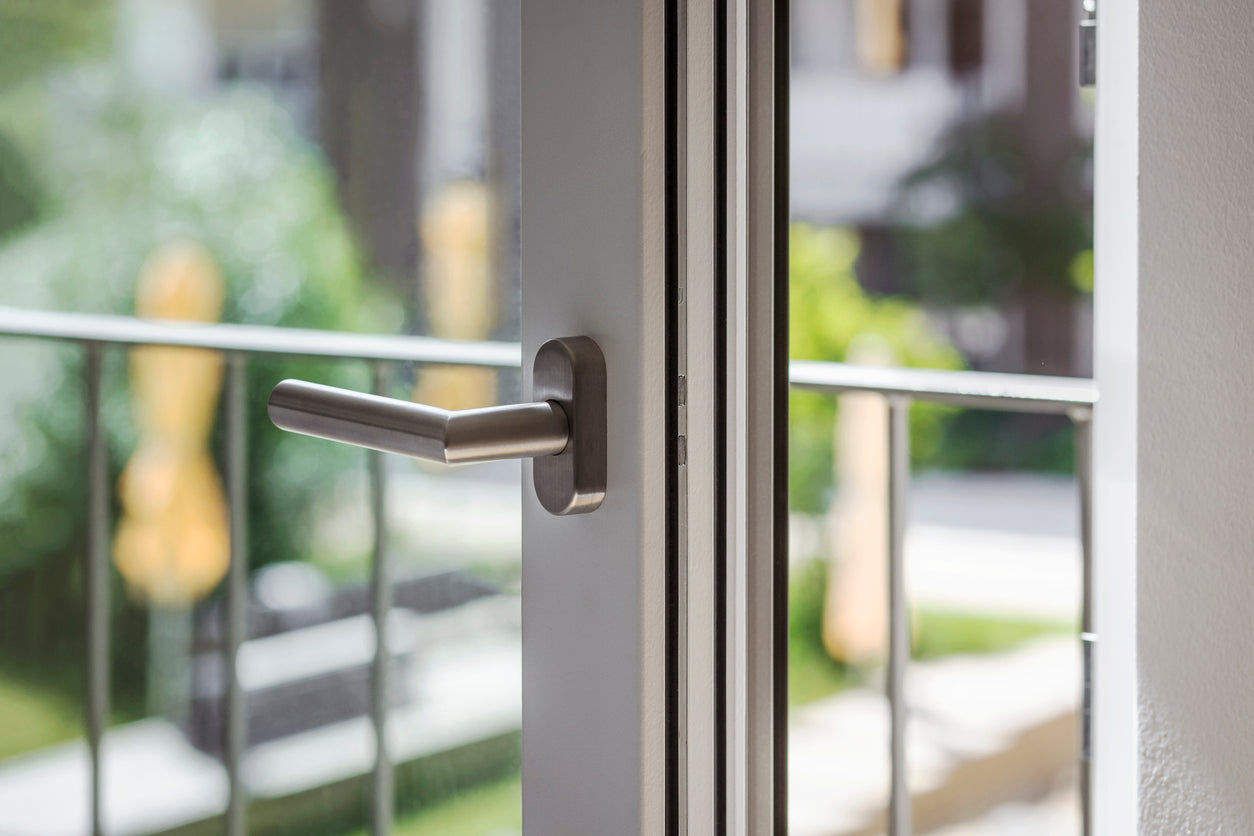 Weisses Fenster mit grauem Griff, Nahaufnahme, im Hintergrund Terrasse und gruenes Umfeld