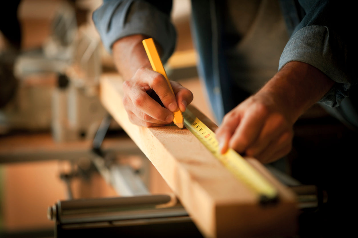 Hand eines Tischlers misst eine Holzlatte, Bleistift und Metermass sichtbar, Werkstattatmosphaere