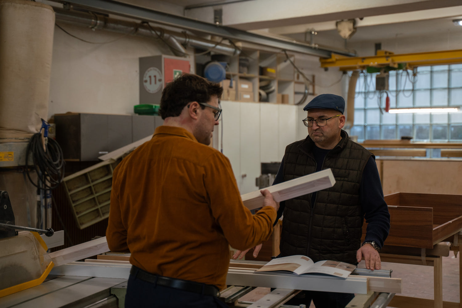 Schreinerund Architekt pruefen eine Holzlatte und einen geoeffneten Katalog in der Werkstatt, warme Arbeitsatmosphaere